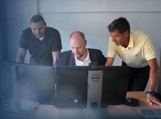 Three development engineers stand together and look at two screens on a desk.
