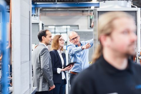 Three employees are standing at a chiller in production, talking and pointing at the technical interior of the chiller.